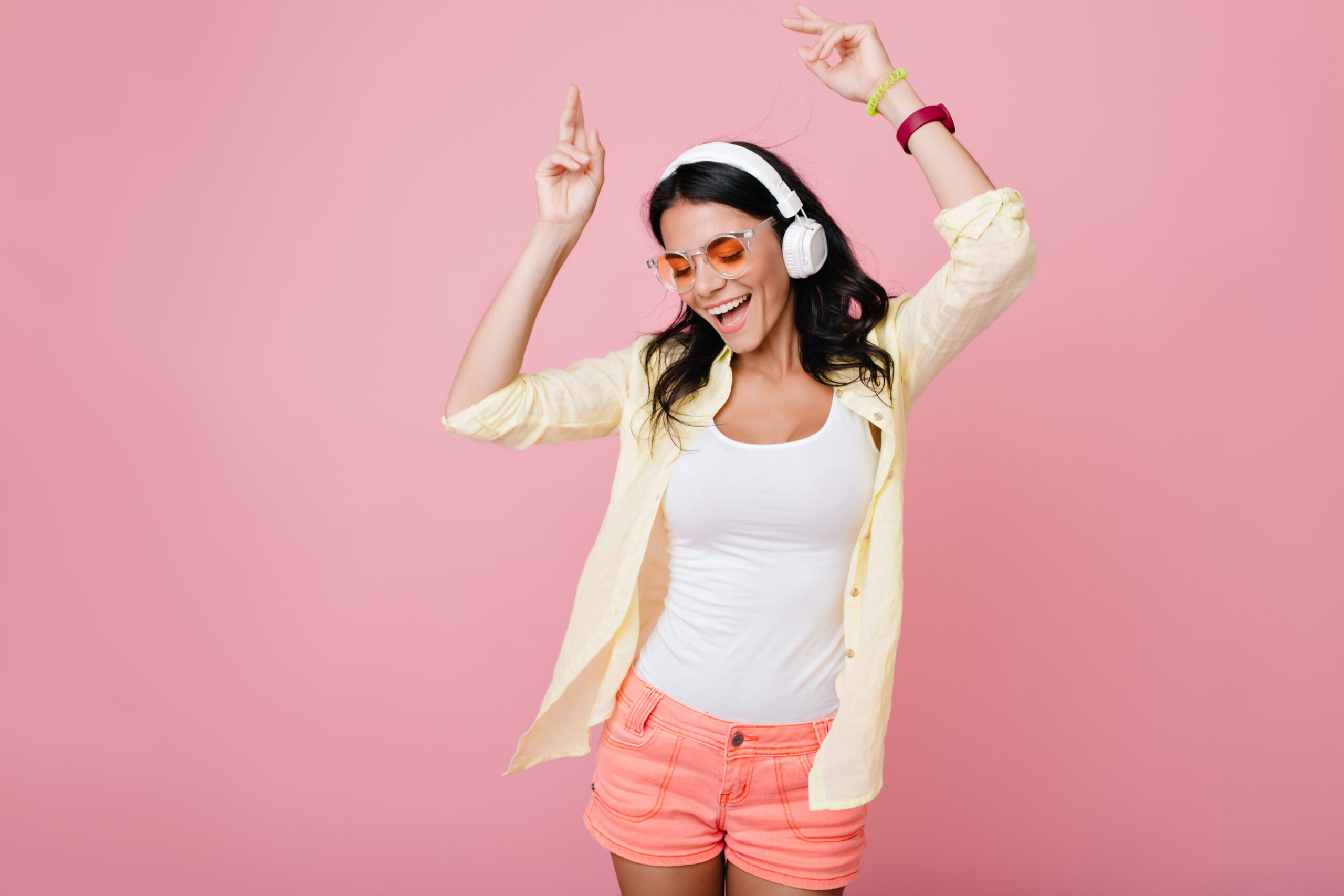 Inspired slim brunette girl in sunglasses funny dancing on pink background and waving hands. Laughing dark-haired young woman in yellow shirt enjoying music in headphones with eyes closed.