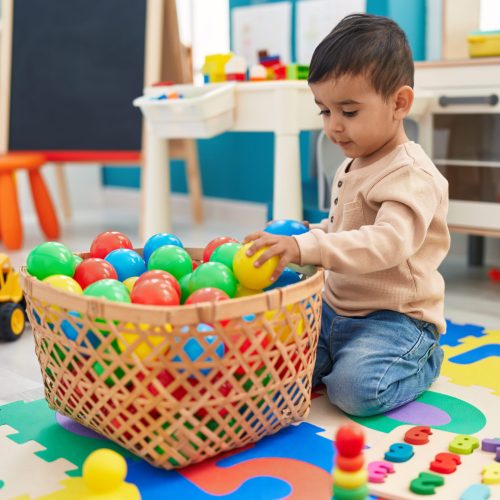 Adorable hispanic toddler playing with balls sitting on floor at kindergarten