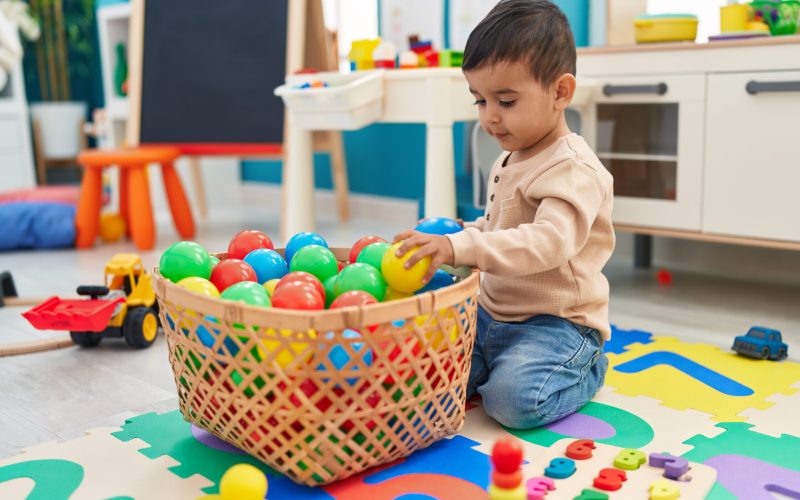 Adorable hispanic toddler playing with balls sitting on floor at kindergarten