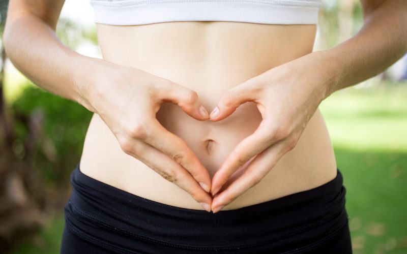 Close-up of female hands shaping heart on belly outdoors. Fitness and dieting concept
