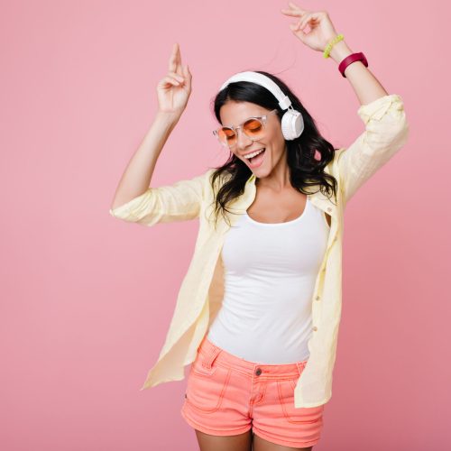 Inspired slim brunette girl in sunglasses funny dancing on pink background and waving hands. Laughing dark-haired young woman in yellow shirt enjoying music in headphones with eyes closed.