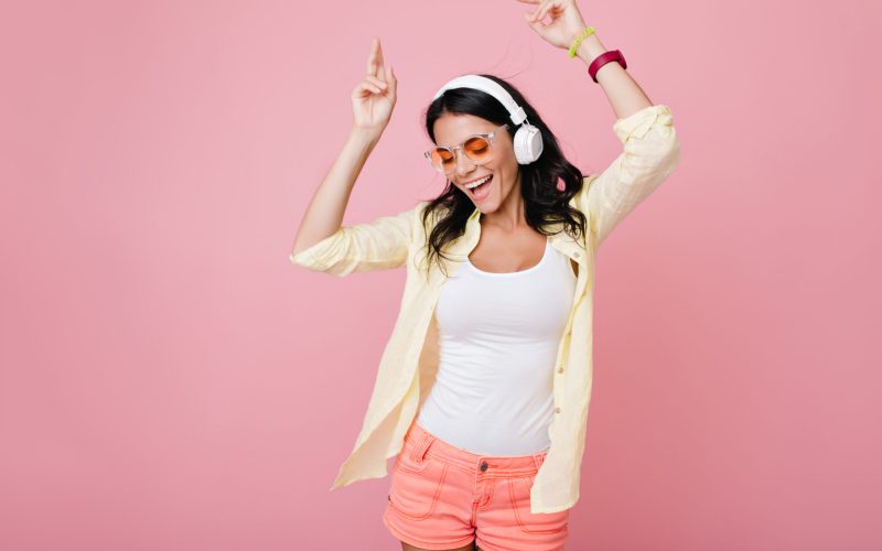 Inspired slim brunette girl in sunglasses funny dancing on pink background and waving hands. Laughing dark-haired young woman in yellow shirt enjoying music in headphones with eyes closed.