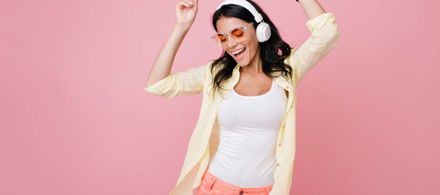 Inspired slim brunette girl in sunglasses funny dancing on pink background and waving hands. Laughing dark-haired young woman in yellow shirt enjoying music in headphones with eyes closed.