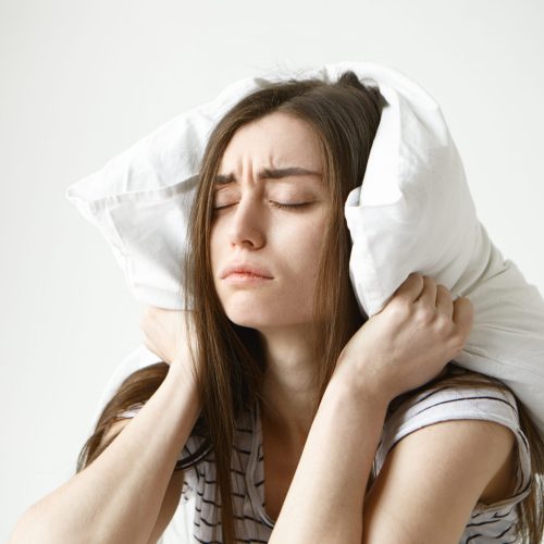 Pretty student girl in striped t-shirt trying to have some sleep, sitting in bedroom with pillow on her head, closing eyes, having tired painful look, irritated with annoying sound. Negative emotions