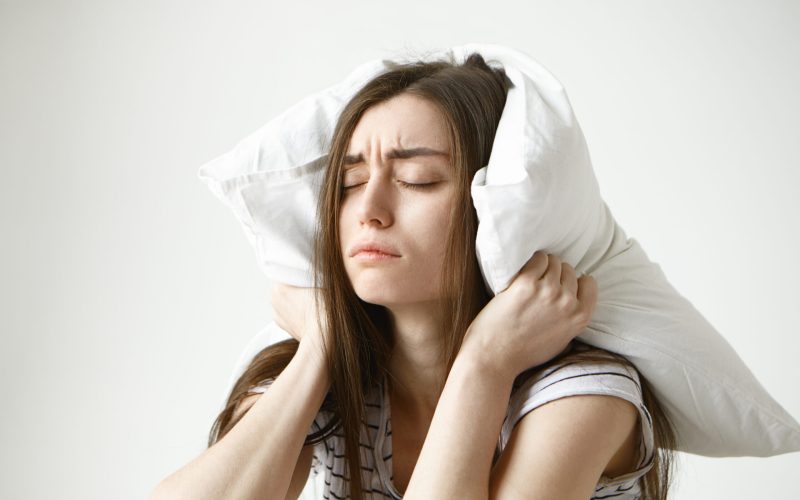 Pretty student girl in striped t-shirt trying to have some sleep, sitting in bedroom with pillow on her head, closing eyes, having tired painful look, irritated with annoying sound. Negative emotions