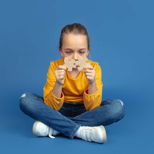 Portrait of sad little girl sitting isolated on blue studio background. How it feels to be autist. Modern problems, new vision of social issues. Concept of autism, childhood, healthcare, medicine.