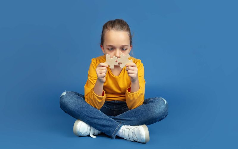 Portrait of sad little girl sitting isolated on blue studio background. How it feels to be autist. Modern problems, new vision of social issues. Concept of autism, childhood, healthcare, medicine.