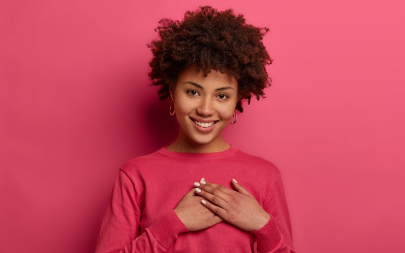 Portrait of touched lovely woman touches heart, makes sign of affection and gratitude, has gentle smile, wears crimson jumper, appreciates nice words, looks gratefully, isolated on pink wall