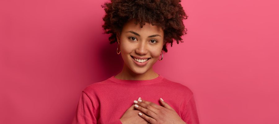 Portrait of touched lovely woman touches heart, makes sign of affection and gratitude, has gentle smile, wears crimson jumper, appreciates nice words, looks gratefully, isolated on pink wall